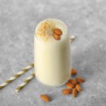 A glass of almond milkshake sits on a table next to two straws and several scattered almonds.